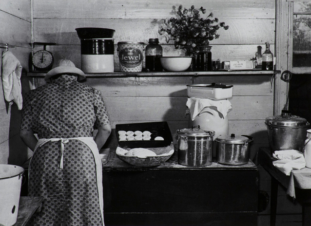 Black and white photograph of back of figure in short sleeve patterned dress with white apron and hat working with dough on a countertop with dough, uncooked biscuits, pots, and pans.