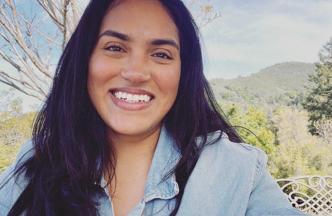 A person smiling outdoors with mountains in the background.