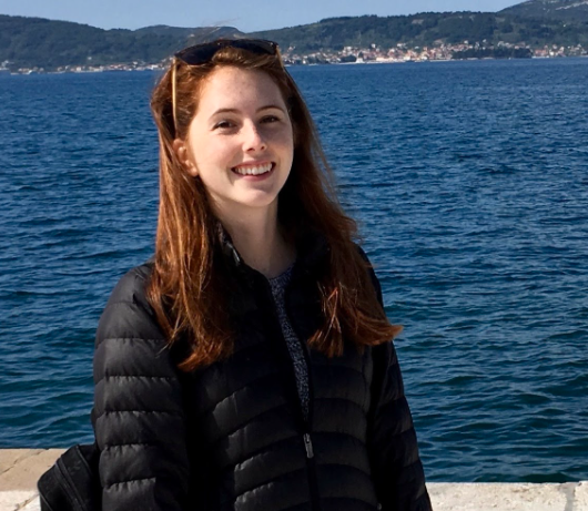 A person smiling by the sea with mountains in the background.