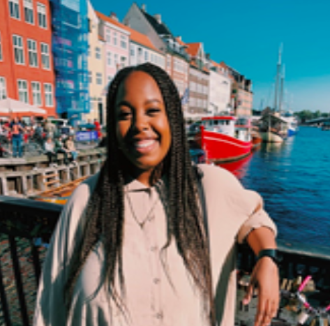 A person smiling in front of colorful buildings by a waterfront.