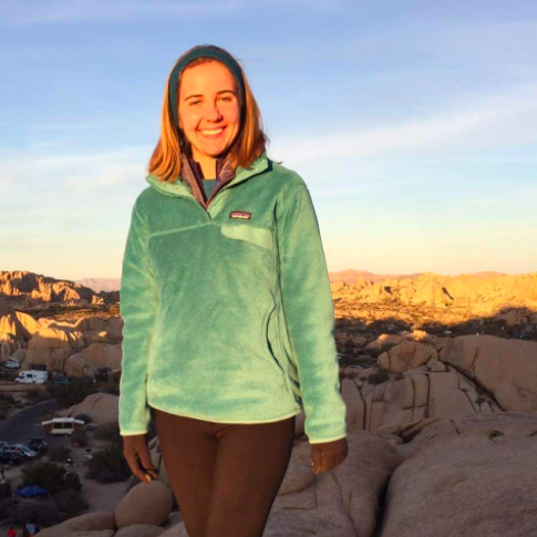 Person in green jacket smiling outdoors at sunset, rocky landscape in background.