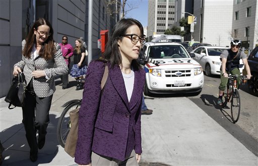 Two women walking on sidewalk in city, with parked cars and people in background.