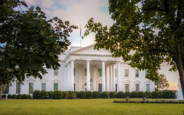 A large white house with columns and many windows. The house sits on a expansive green lawn with a large circular fountain and is framed by two green leafy trees.