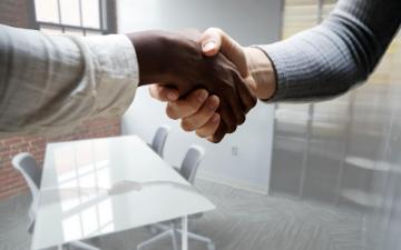 Two hands reaching across a table in a handshake. The reflection of their hands can be seen in the glass tabletop. image link to story