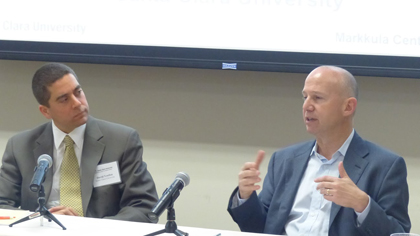 Two men in suits discussing at a conference panel.