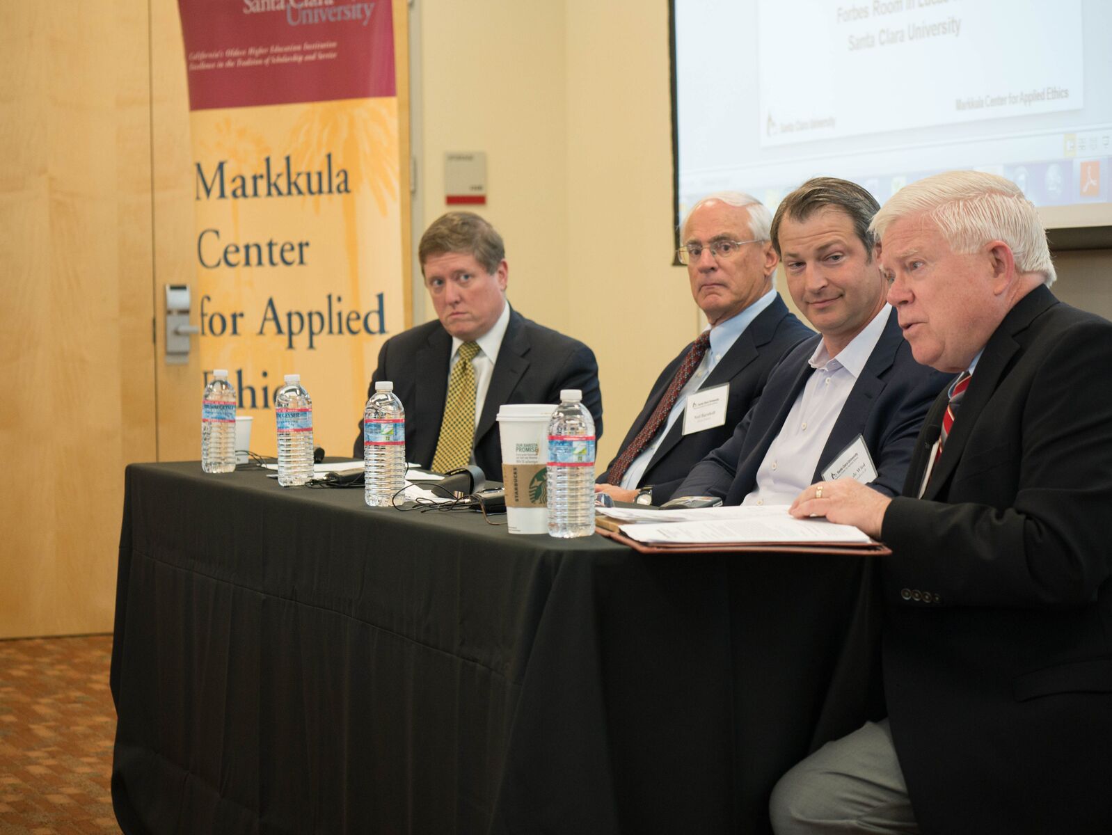 Four men seated at a panel discussion with a banner in the background.