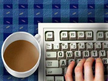 A hand typing on a keyboard next to a cup of coffee.