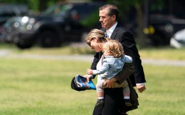 Hunter Biden carrying a young child while walking outdoors in a grassy area.