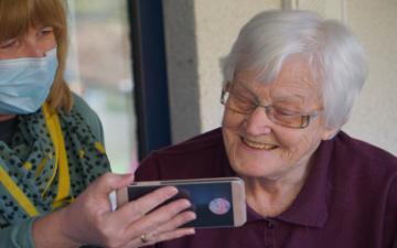 A senior person looking at a smartphone, assisted by a person wearing a mask.