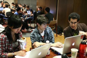 People collaborating at a table with laptops and beverages in a busy room.