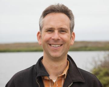 A person standing near a body of water, smiling.