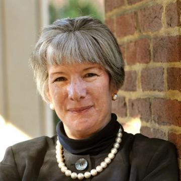 A person wearing a black outfit and pearl necklace, leaning against a brick wall.