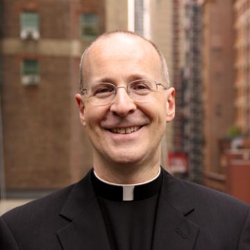 A smiling man in a clerical collar and black attire, standing outdoors.