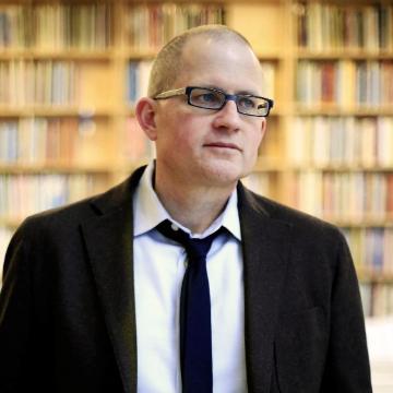 A person wearing glasses, a suit, and a tie in front of a bookshelf.