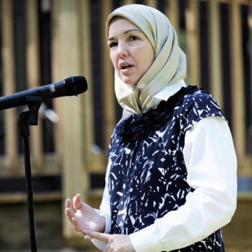 A woman speaking at a microphone, wearing a headscarf and patterned outfit.