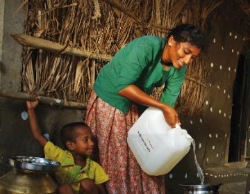 A woman pours water from a large container by a hut. image link to story