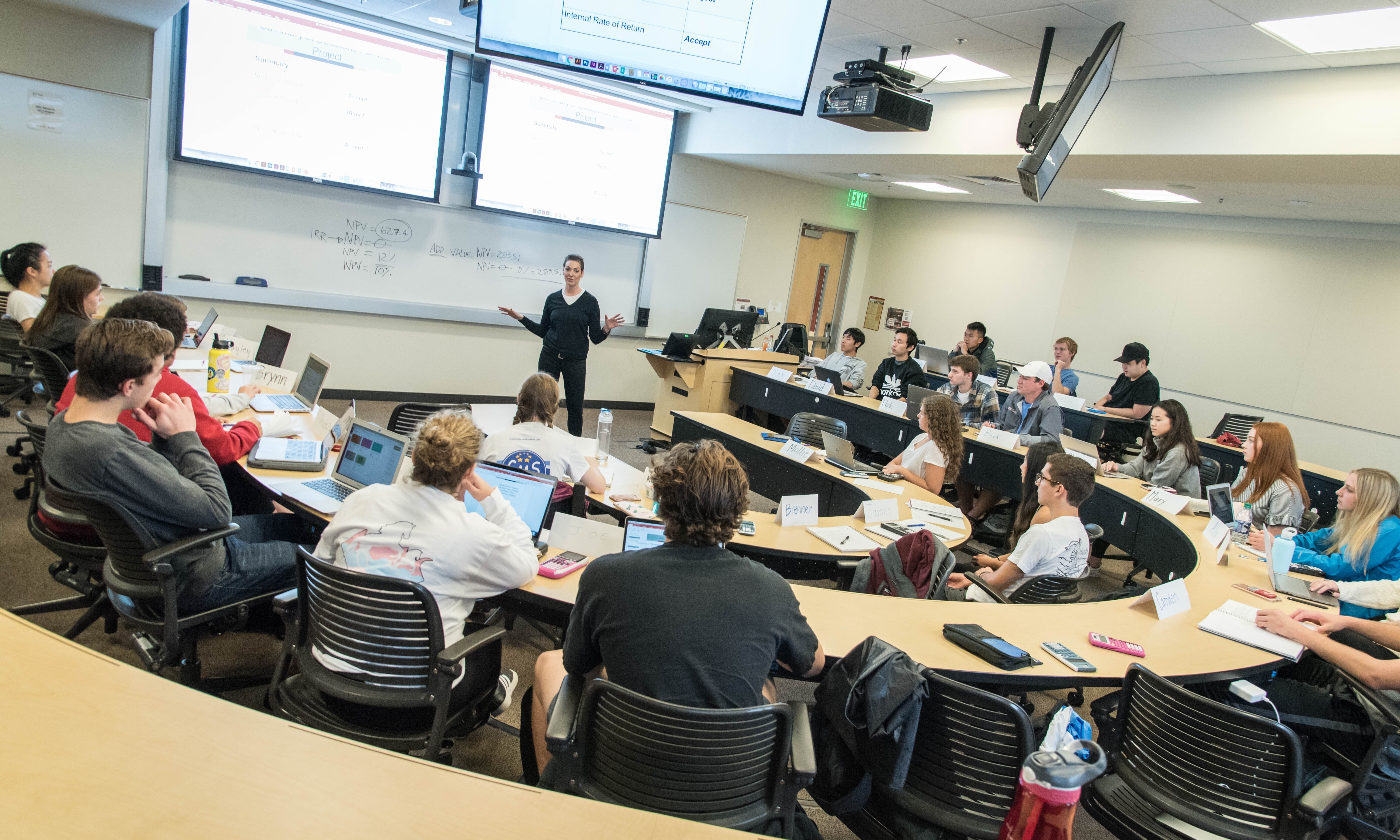 Alt text: Professor lecturing to a full classroom of students with multiple screens displaying information.