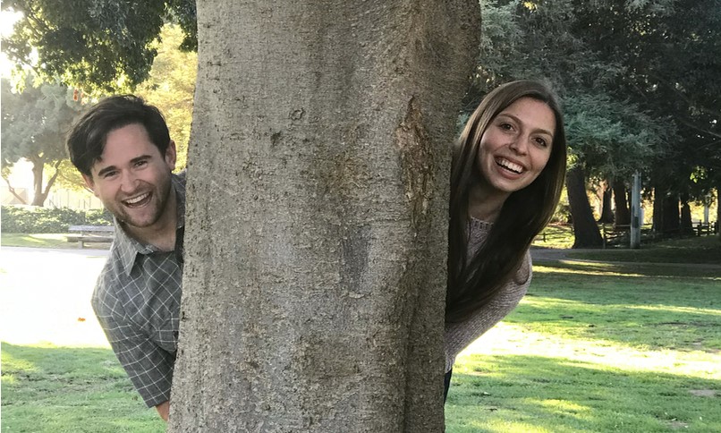 Two people peeking from behind a tree in a park.