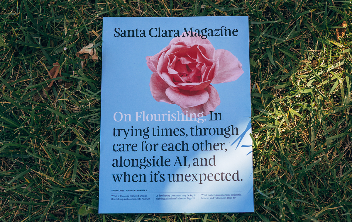 Spring 2026 issue of Santa Clara Magazine laying on the grass. Cover features blue background with pink flower, reads, On flourishing. In trying times, through care for each other, alongside AI, and when its unexpected.