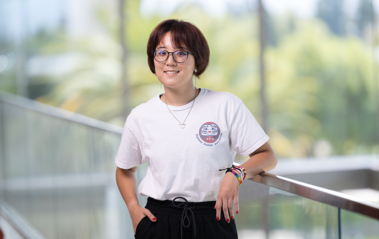 A student wearing glasses and a shirt from the Japanese Student Association leans on a glass balcony railing with open space, tall windows, and trees in the background.