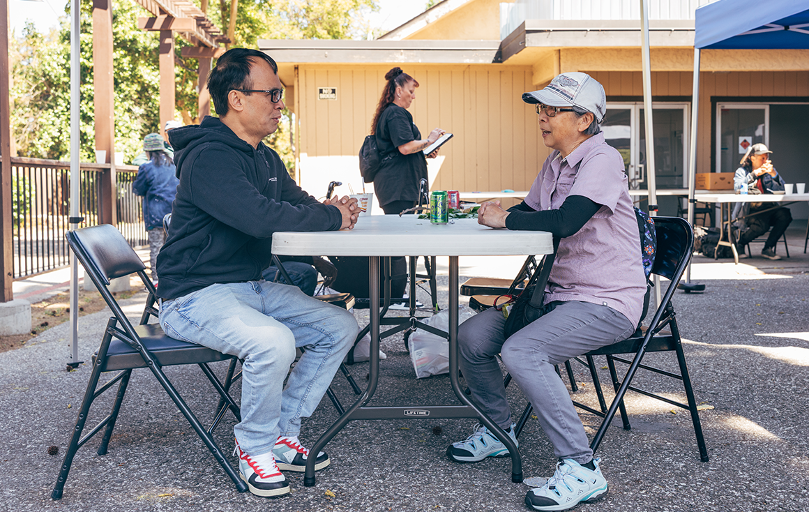 A man and woman sit facing each other at a foldable table in a outdoor courtyard.