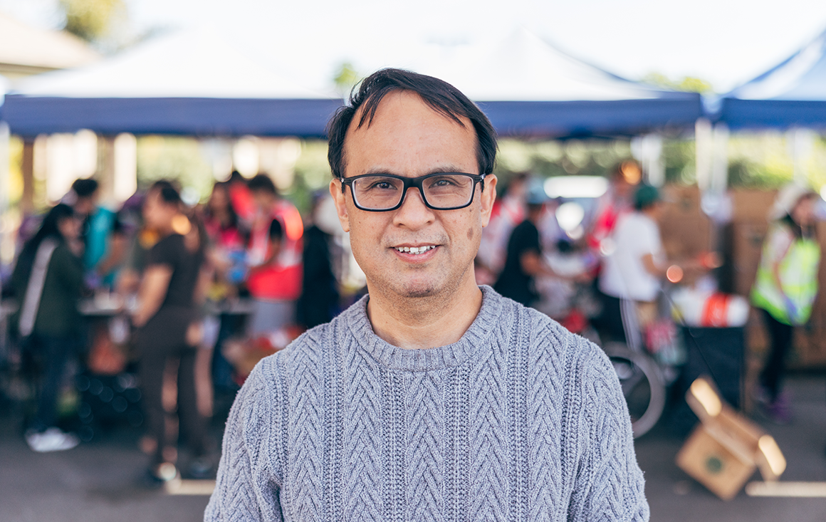 A man in a grey cardigan stands with a line of food bank volunteers out of focus in the background.