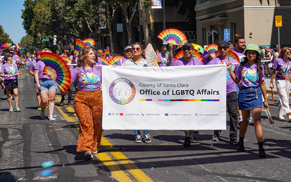 A group of people from the County of Santa Clara Office of LGBTQ Affairs in purple shirts and rainbow accessories marching in a Pride parade.