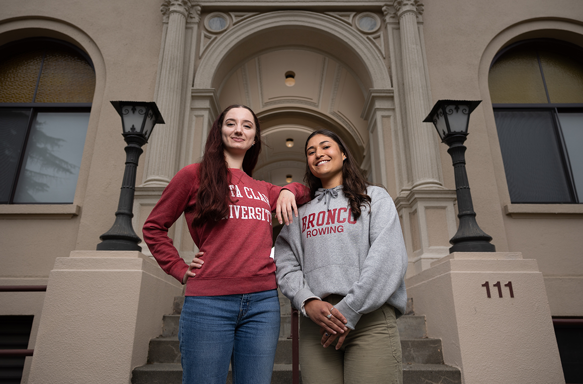 Two students stand at the foot of a staircase by a building's arched entrance.