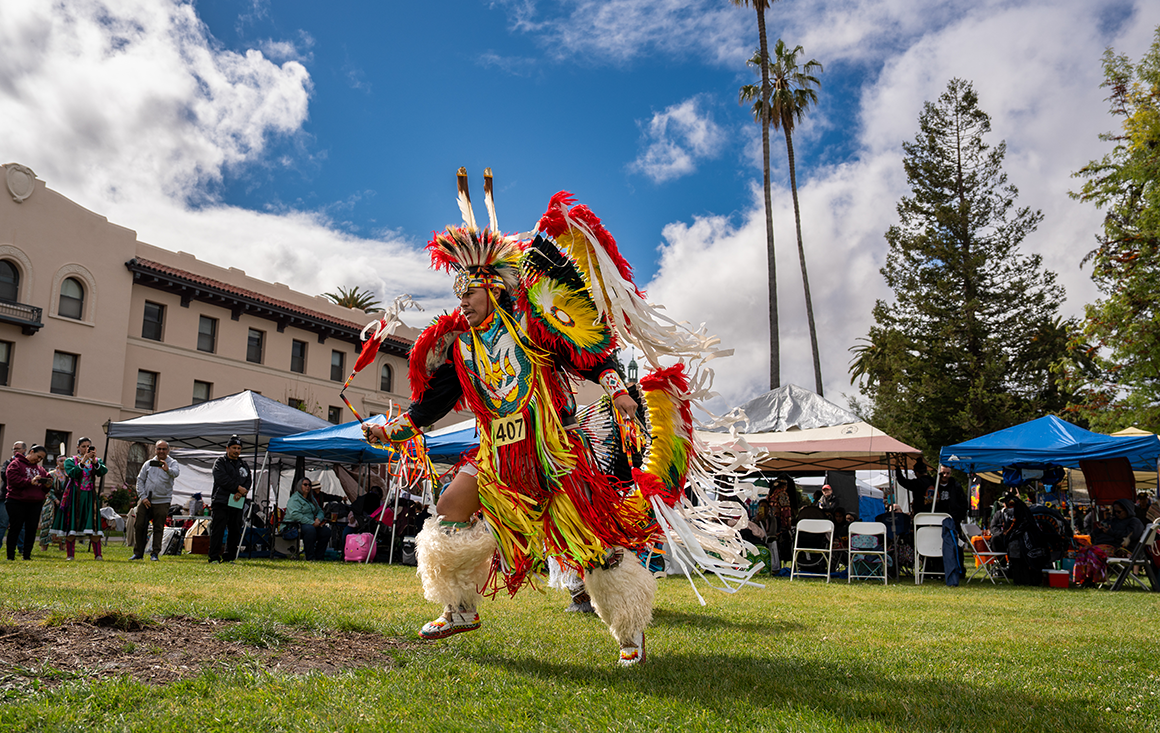 An indigenous man in red tribal regalia with ribbons and feathers dances on a lawn at Santa Clara University.