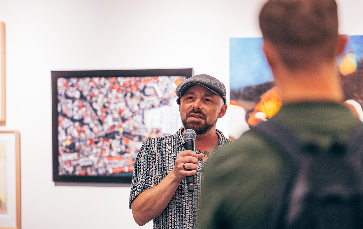 A person in a flat cap speaks into a microphone to a crowd in an art gallery.