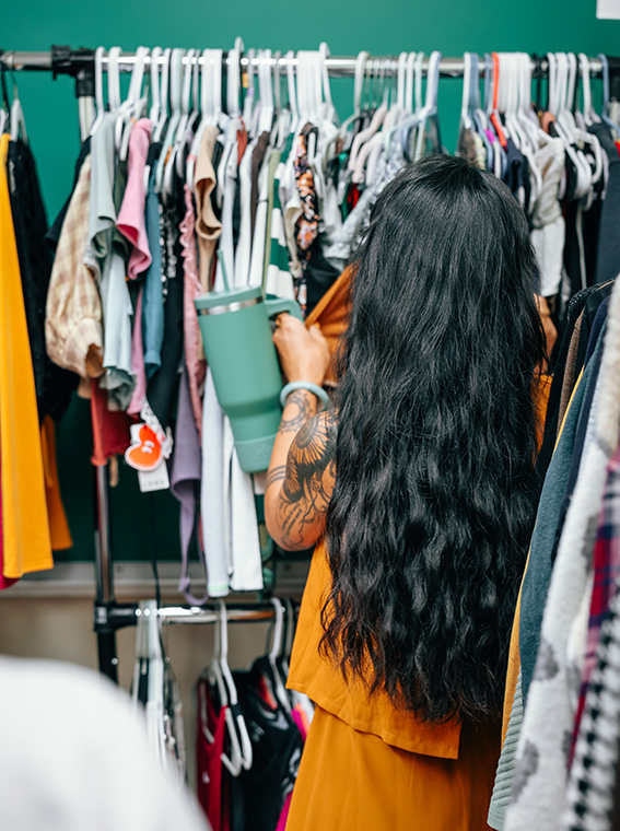 Photo of a woman shopping in a rack of clothes, holding a large light blue cup.