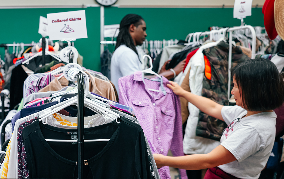 Woman standing in foreground next to clothes rack holds up purple shirt. Man in background looks through different rack of clothes.