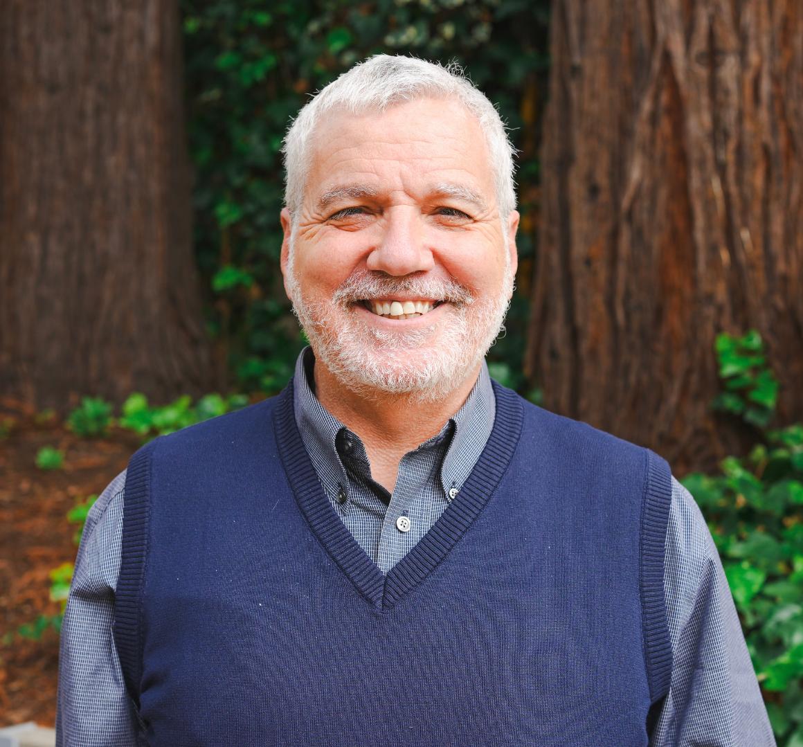 An older man wearing a navy blue sweater vest smiles outside