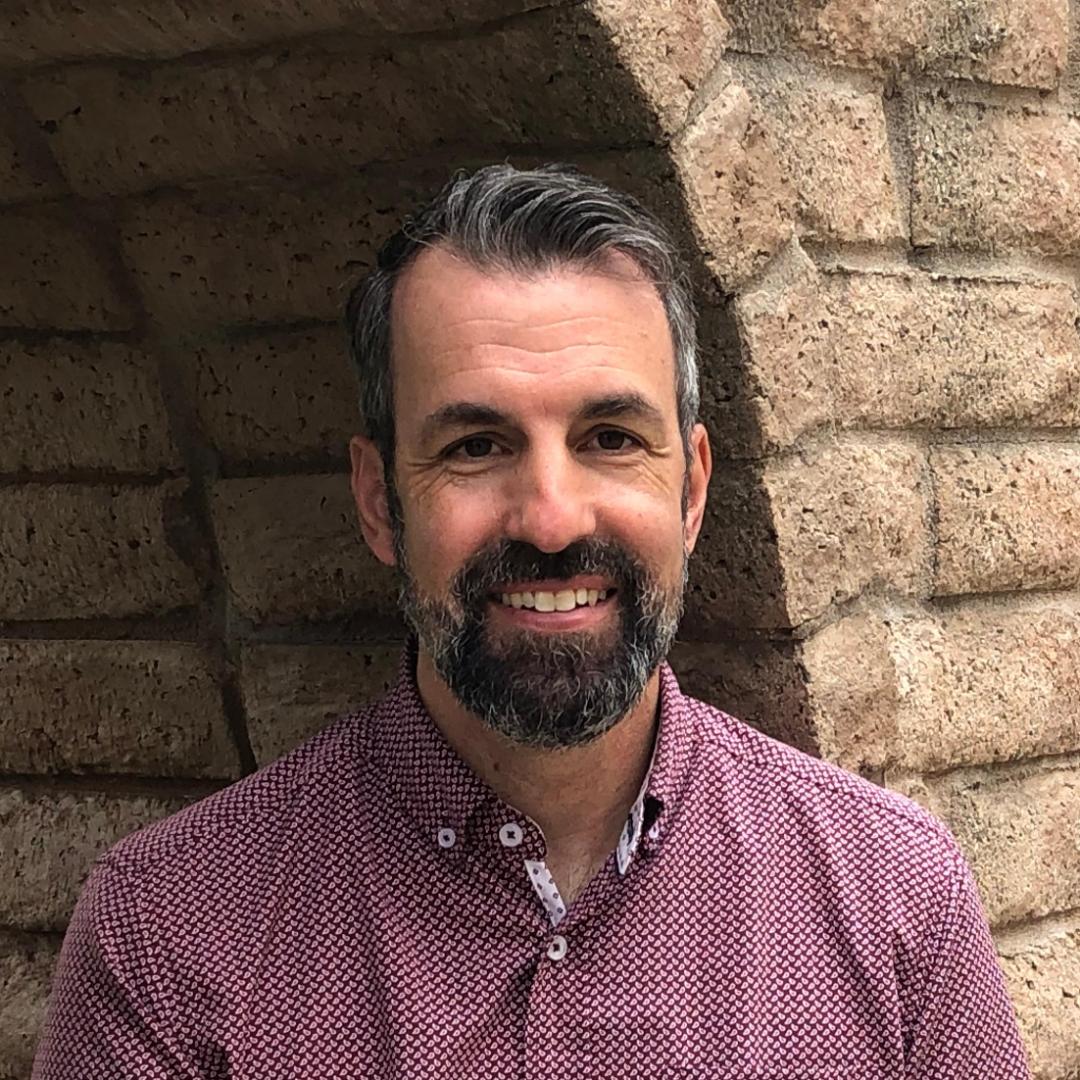 Man in a maroon shirt smiling in front of a stone arch.