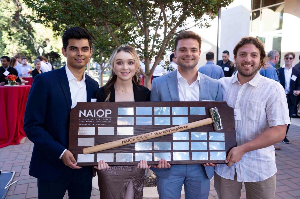 Four people holding a large check outside with trees in the background.