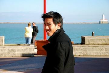 A person smiles near a waterfront with people in the background.