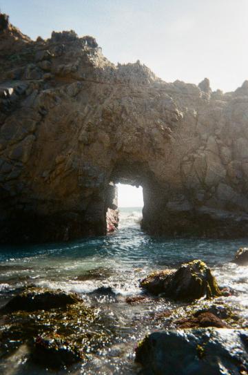 Sunlight shines through coastal rock arch near the ocean with boulders in the foreground.