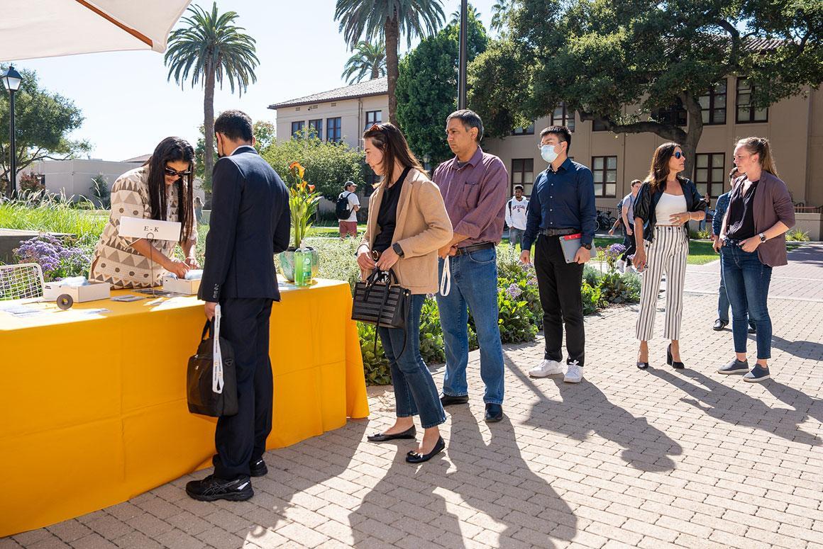 People at an outdoor event, standing in line by a yellow table.