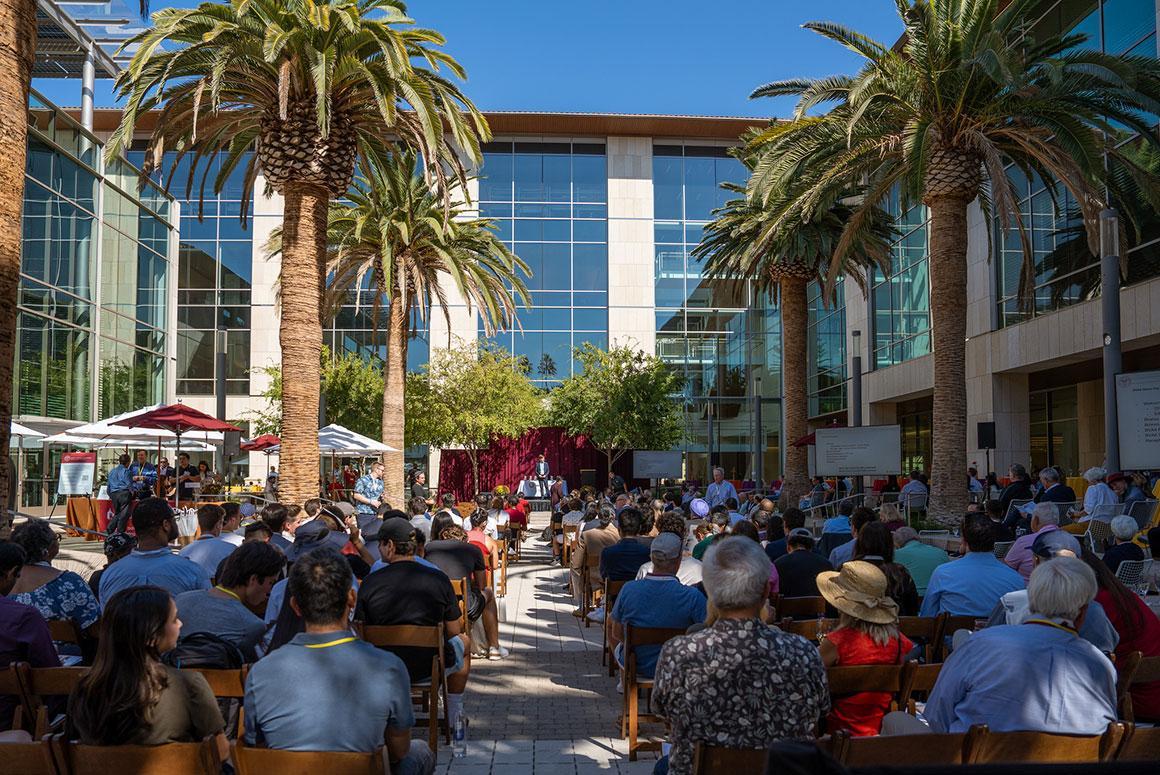 Outdoor event with people sitting and listening to a speaker. Palm trees frame the scene.