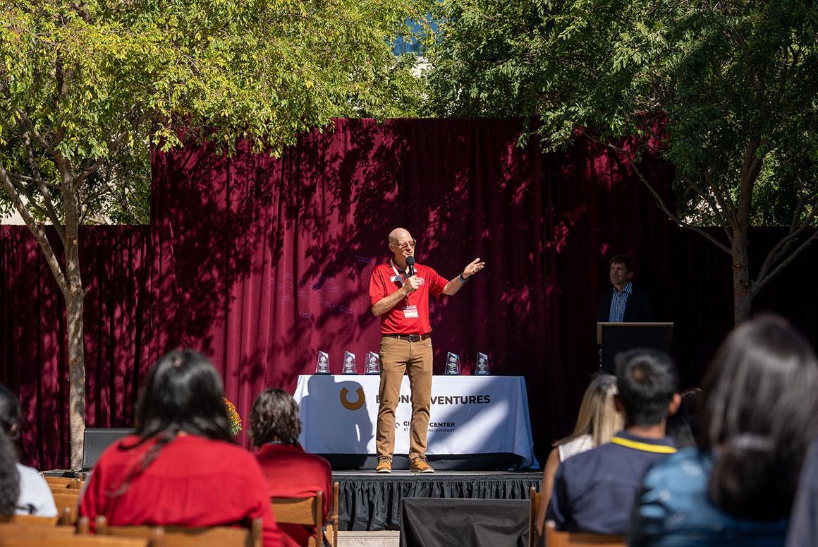 Person speaking on stage with audience at outdoor event titled 