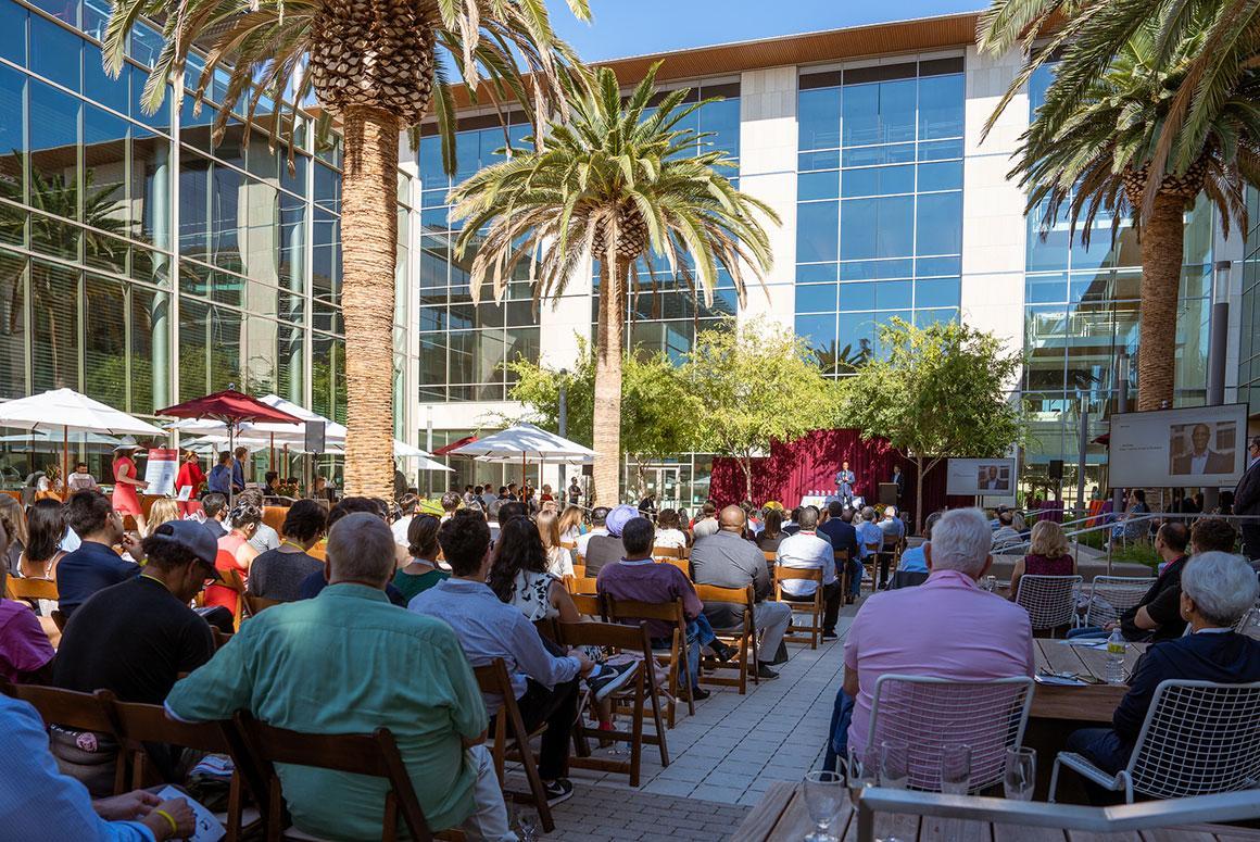 People seated outdoors at an event with a modern building background.