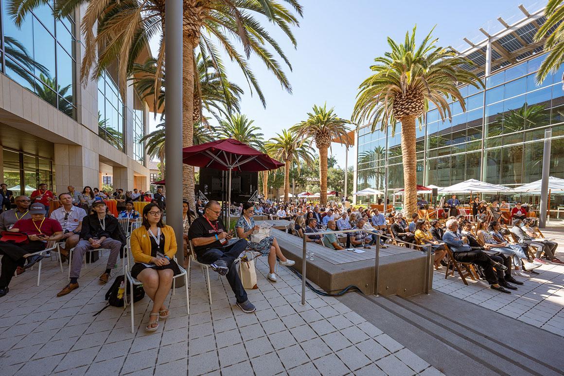 A large outdoor gathering with people attending an event under palm trees.