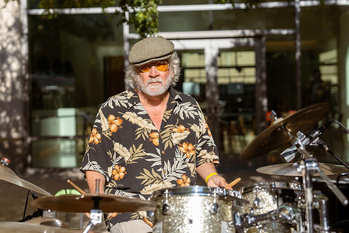 A man in a floral shirt plays the drums outdoors.