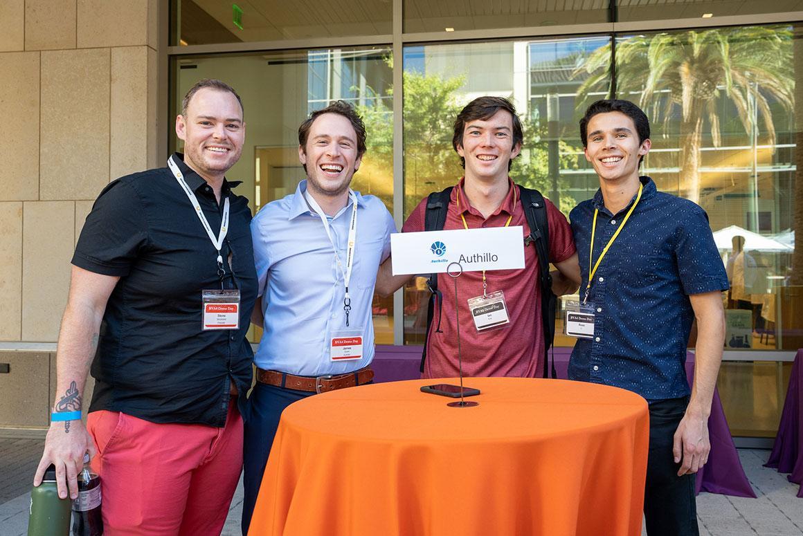 Four people at an event, standing behind an orange-covered table.