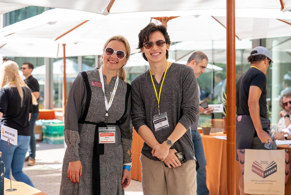 Two people smiling at an outdoor event with name tags on lanyards.