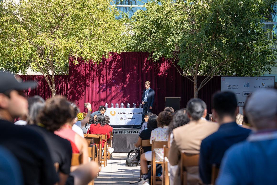 A person presenting on stage before an outdoor audience.