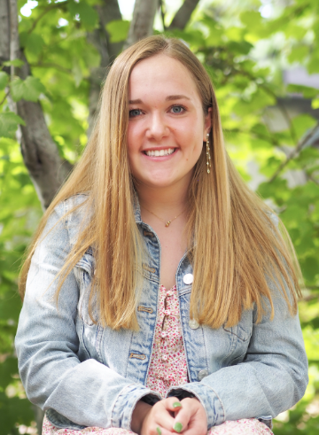 A person smiling, wearing a denim jacket, with a green leafy background.