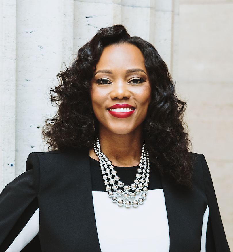 A person smiling, wearing a black and white outfit with a statement necklace.