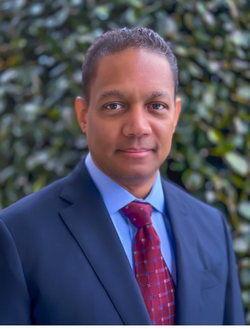 A man in a suit smiling in front of a leafy background.