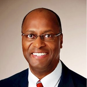 A person wearing glasses, a suit, and a tie smiles.